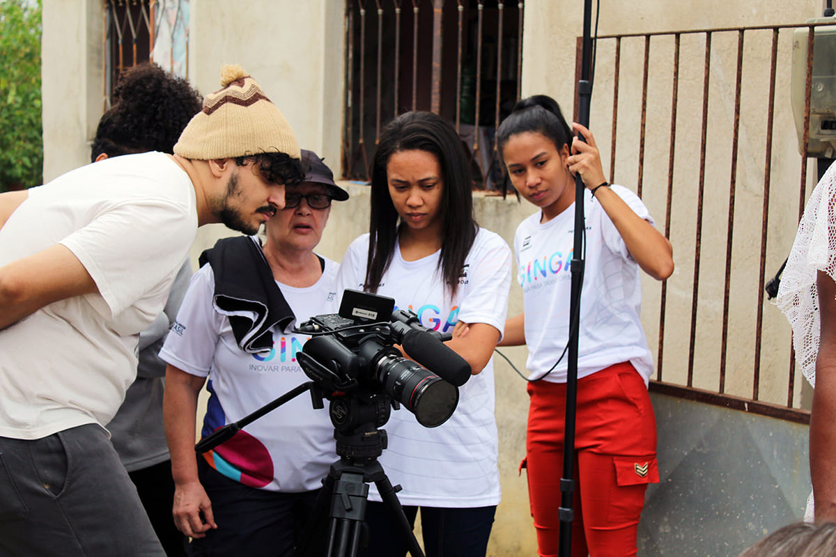 Cinco pessoas durante filmagem em área externa analisam imagens de câmera de vídeo. Algumas usam a camiseta do projeto Ginga, da instituição sociocultural Cinema Nosso, responsável pelo curso de Roteiro e Cinema para Direitos Humanos.