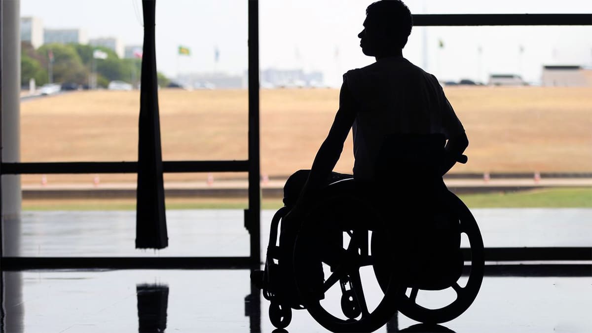 Silhueta de pessoa em cadeira de rodas - equipamento que pode ser adquirido com nova linha de crédito para PcDs -, parada em frente a uma grande janela.