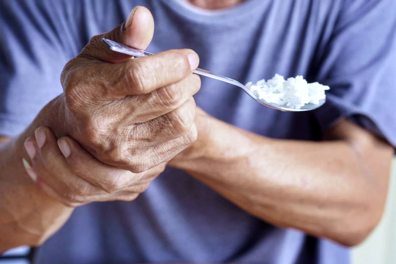 Pessoa tenta estabilizar a mão para se alimentar, com tremores da doença de Parkinson, segurando o próprio punho, enquanto leva colher à boca. Estima-se que o Parkinson afeta 10 milhões pessoas.