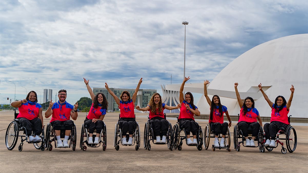 Integrantes da Cia Street Cadeirantes em fila, lado a lado, acenando e celebrando. Ao fundo é possível enxergar o Senado Federal e a Catedral Metropolitana, em Brasília (DF).