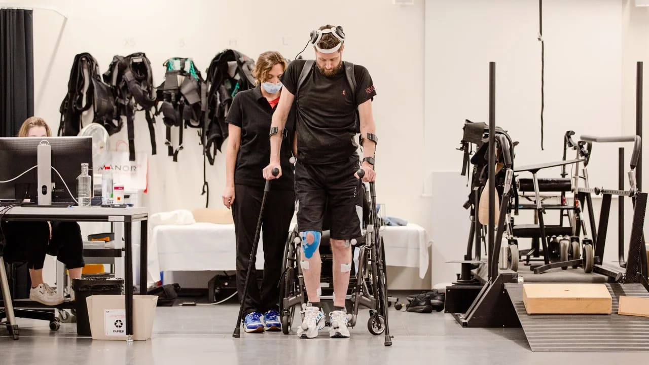 Imagem mostra como um tetraplégico volta a andar após implante experimental. Na fotografia, o paciente está de frente e se apoia em muletas, Atrás dele, uma cientista no Hospital Universitário de Lausanne, EPFL, está em pé observando a marcha.