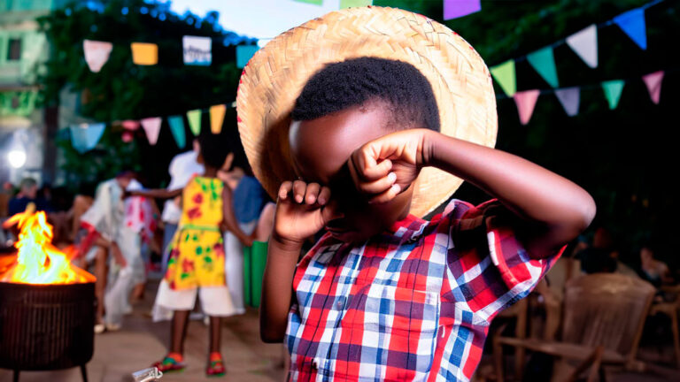 Uma criança autista em festa junina. Está com as mãos fechadas, tampando o rosto, demonstrando estar incomodada. Ela é negra, usa chapéu de palha e camisa xadrez. Ao fundo a imagem desfocada com bandeirinhas, fogueira, mesas e outras pessoas.