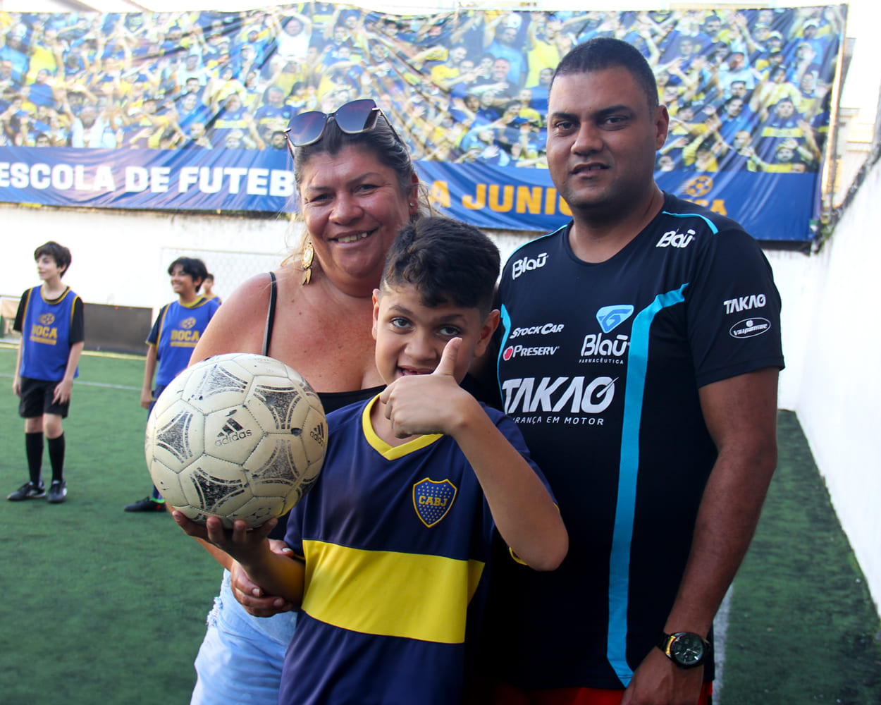 O jovem Enzo está segurando a bola e acenando com um sinal de positivo. Ele usa o uniforme do Club Atlético Boca Juniors, nas cores azul e amarelo, e sorri. Atrás dele está a sua mãe, à esquerda, e o seu pai, à direita. Em segundo plano, no campo de futebol, há outros alunos da escolinha.