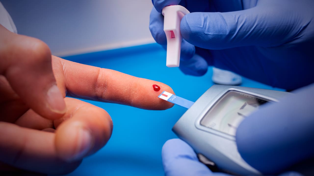 Foto de exame de prevenção do diabetes, mostra as mãos de profissional da saúde com luvas, segurando o medidor de glicose enquanto colhe sangue do dedo indicador de paciente.