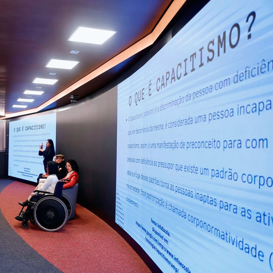 Sala Victor Civita, com detalhe da tela de projeção com a pergunta: O que é Capacitismo?, as pessoas palestrantes, mediadora e intérprete de Libras.