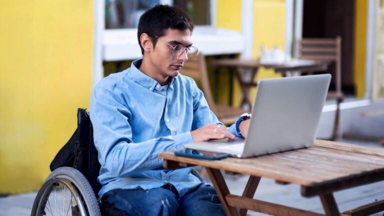 Jovem em cadeira de rodas, usando um laptop em uma mesa de madeira. Ele é branco, tem cabelo curto preto e usa óculos de grau. Ilustra o bootcamp Inclusivo de Front End com bolsas 100% gratuitas para pessoas com deficiência.