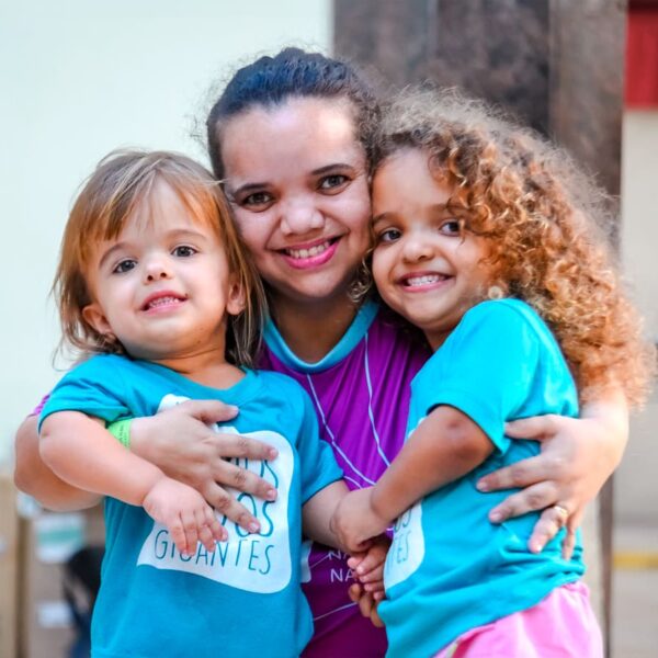 Mulher com nanismo abraçada a duas crianças com a mesma condição, todas sorrindo. Ilustra o mapa do nanismo no Brasil, usam camisetas das organizações Nanismo Brasil e Somos Todos Gigantes.