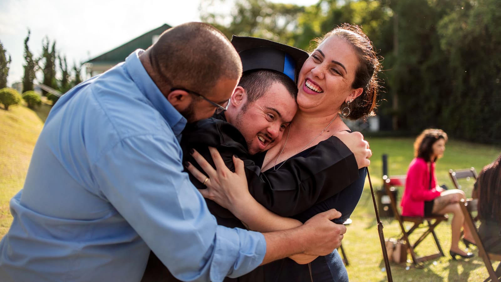 Três pessoas se abraçam em celebração de formatura, entre elas, um jovem com deficiência usando beca e capelo. Ilustra a deficiência na mídia, com a inclusão interseccional na narrativa da deficiência na web.
