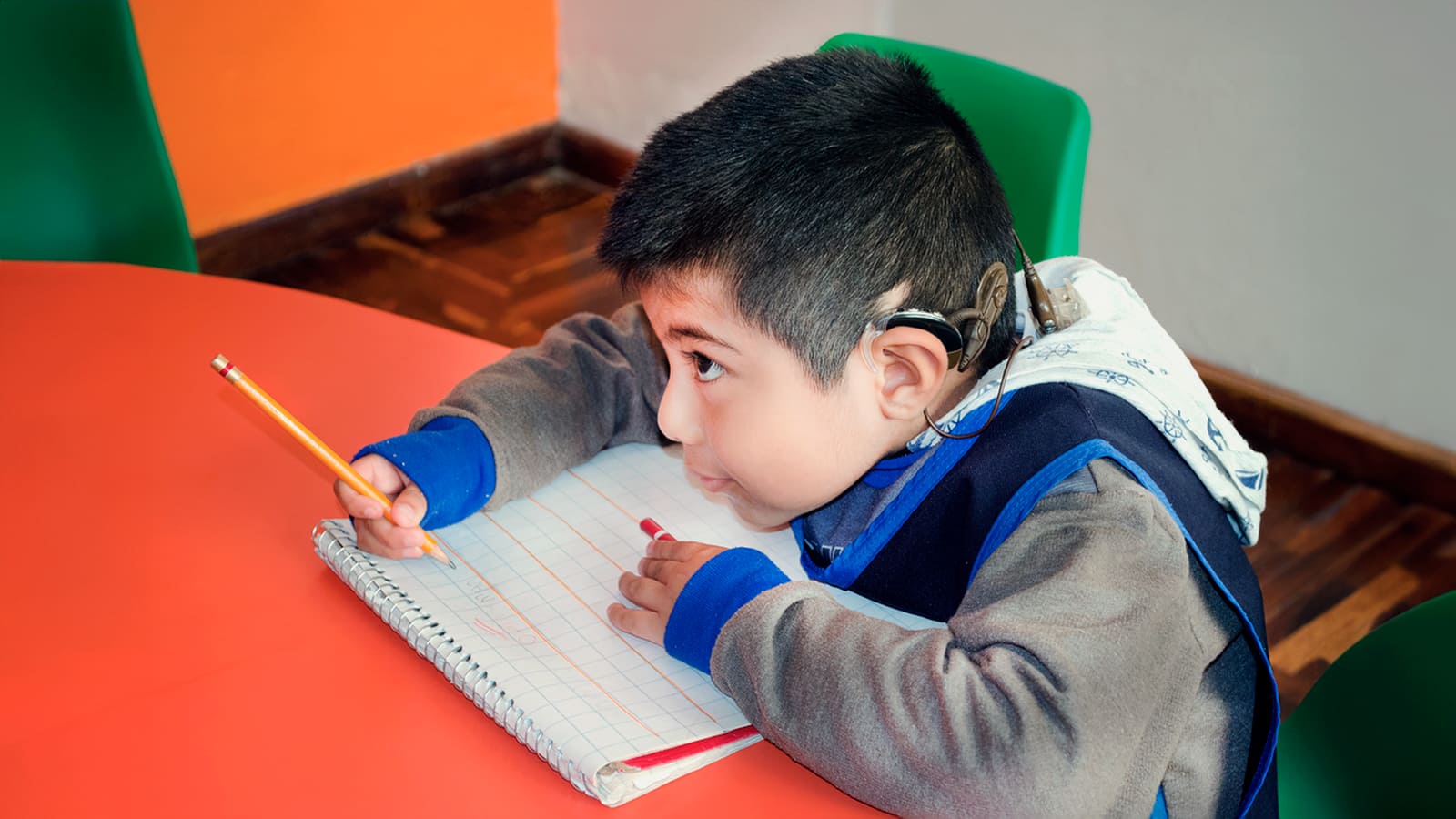 Foto de criança surda implantada coclear, escrevendo no caderno sobre uma mesa laranja. A imagem ilustra o direito à educação.