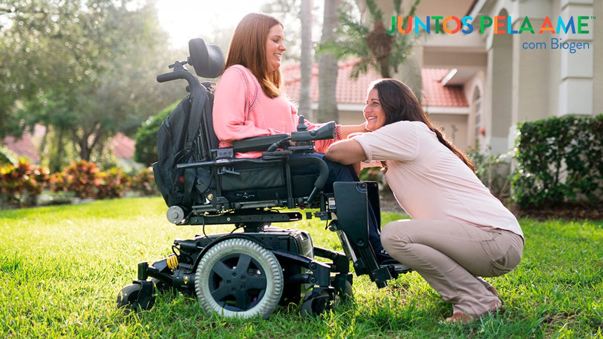 Mulher com atrofia muscular espinhal sentada em cadeira de rodas e outra mulher agachada à sua frente.