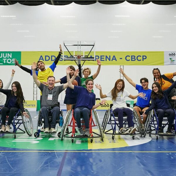 Jovens, adolescentes e adultos estão lado a lado, em cadeiras de rodas preparadas para prática de esportes. Atrás há pessoas em pé e todas estão acenando com as mãos e sorrindo, na quadra poliesportiva da Reatech, na edição de 2022.