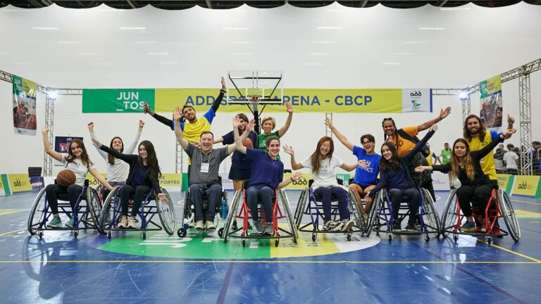Jovens, adolescentes e adultos estão lado a lado, em cadeiras de rodas preparadas para prática de esportes. Atrás há pessoas em pé e todas estão acenando com as mãos e sorrindo, na quadra poliesportiva da Reatech, na edição de 2022.