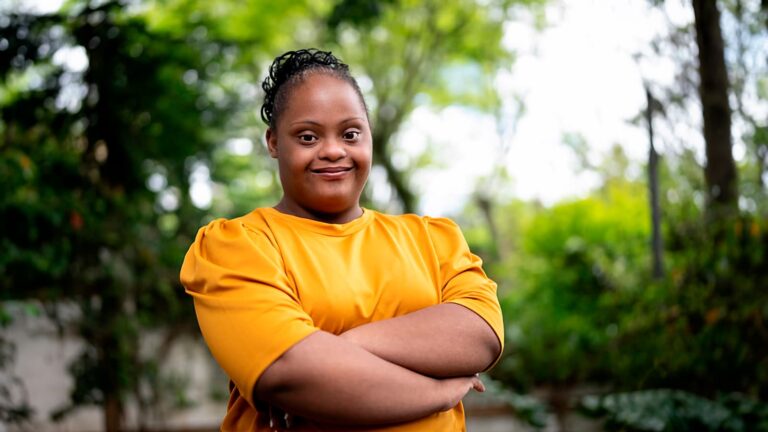 Mulher negra com síndrome de Down, em pé em um jardim durante o dia. Ela veste camiseta amarela e tem os braços cruzados. O fundo é composto por árvores e plantas. Ilustra o lançamento do 1º Observatório da Deficiência Intelectual do Estado de São Paulo.