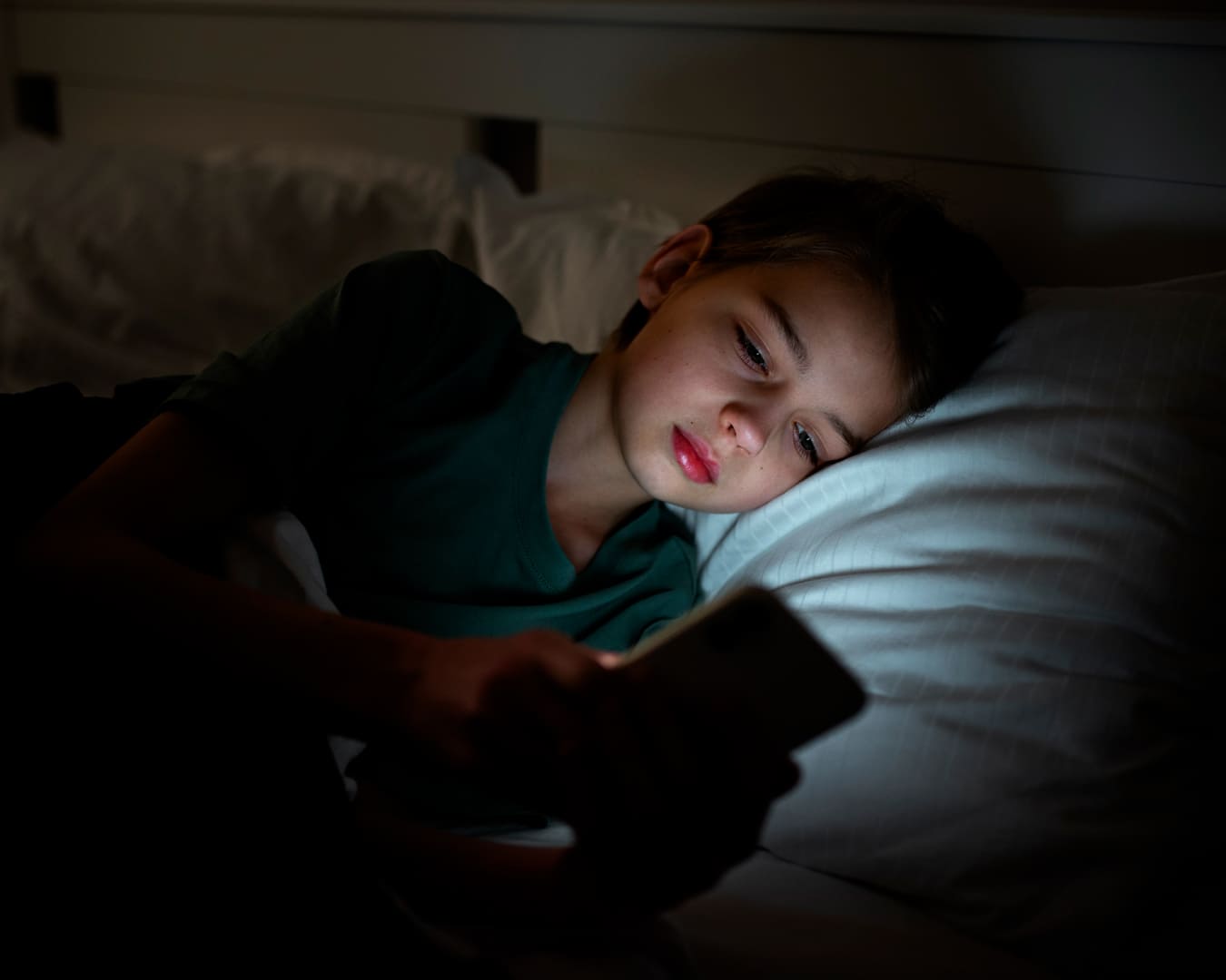 Adolescente deitado na cama e segurando um telefone em um quarto escuro.