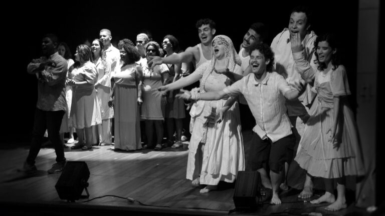 Foto em preto e branco da Orquestra Brasileira de Cantores Cegos, que estreia no Sesc, durante ensaio com a Cia Poéticas Contemporânea. São pessoas de diferentes idades, gênero e raça, com deficiência visual.