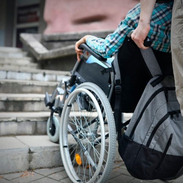 Pessoa em cadeira de rodas sendo conduzida por outra pessoa. Na imagem, elas estão paradas em frente a um conjunto de escadas, em alusão a petição “Diga Não ao PL 3835”. Acessibilidade em Hotéis é Lei.