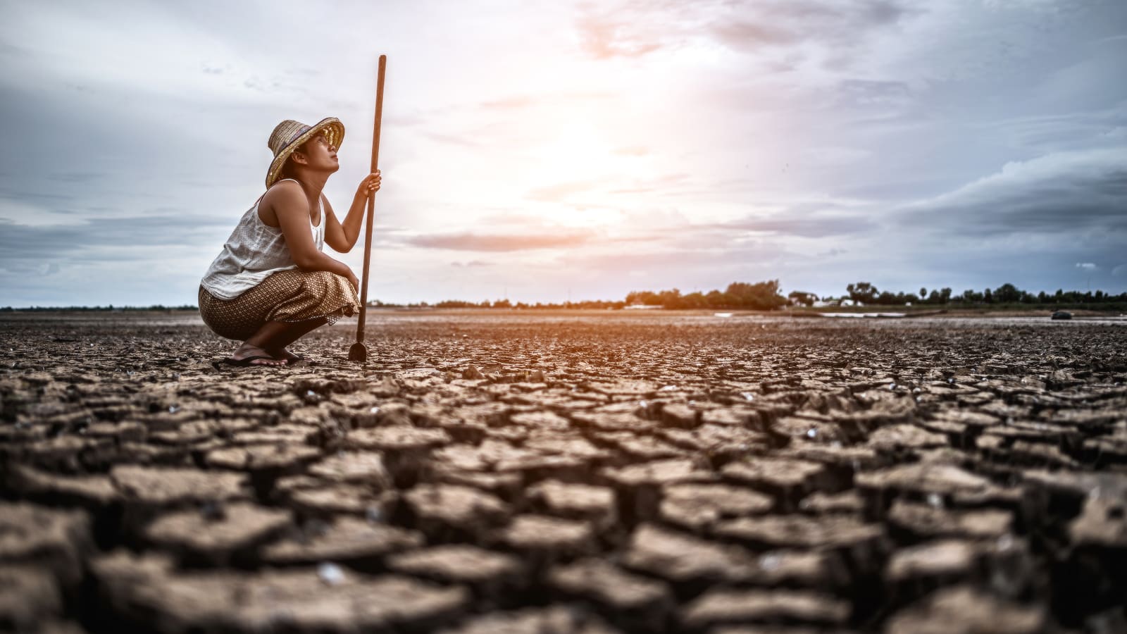 Foto em ambiente externo mostra uma pessoa sentada de cócoras em um solo com erosão, retratando as mudanças climáticas e seus impactos na saúde. A pessoa usa chapéu de palha e olha para o céu.