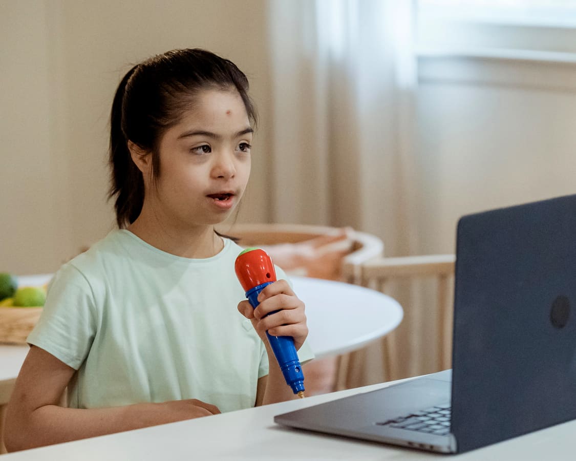 Criança com T21 brincando de karaokê, com uma microfone vernelho e um laptop.