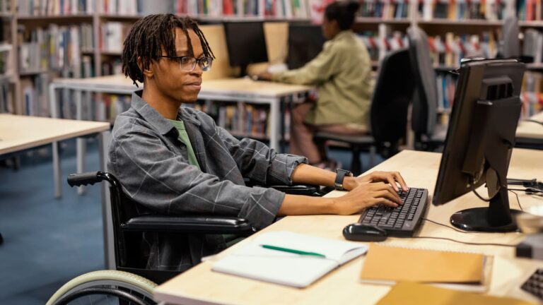 Pessoa em cadeira de rodas usando um computador em uma biblioteca. Programa Desenvolve 2024.