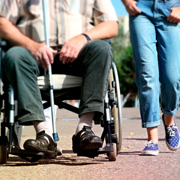 Pessoa em uma cadeira de rodas e outra pessoa caminhando lado a lado em uma calçada.