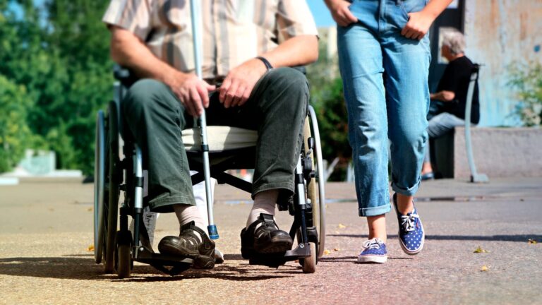 Pessoa em uma cadeira de rodas e outra pessoa caminhando lado a lado em uma calçada.