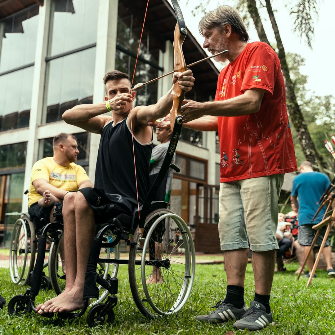 Uma pessoa em uma cadeira de rodas praticando arco e flecha com assistência de um instrutor no Camping Acessível.