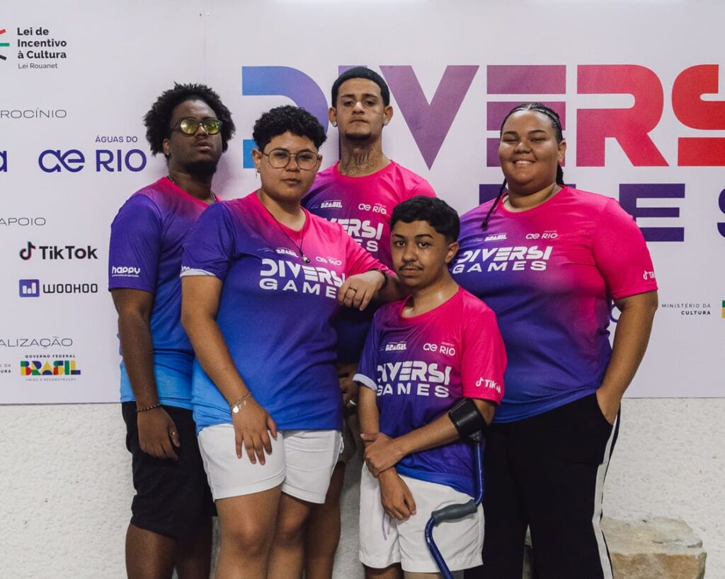 Grupo de pessoas usando camisetas do projeto ‘DiversiGames’ em frente a um pano de fundo da marca, patrocinadores e apoio.