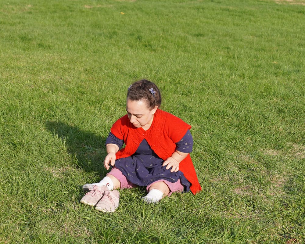 Natalia Grace, uma mulher com Displasia Espondiloepifisária, sentada em um campo gramado, vestindo roupas coloridas; uma cena do documentário ‘O Curioso Caso de Natalia Grace’.