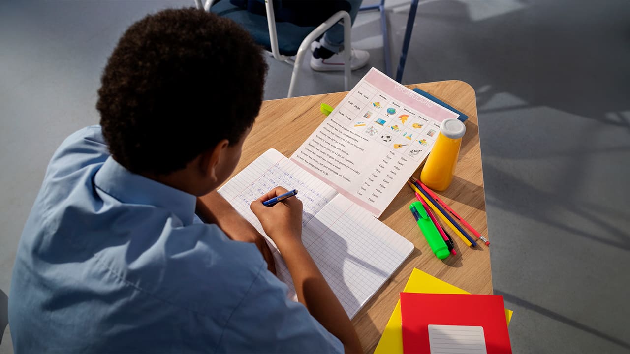 Estudante focado escrevendo em um caderno, com diversos materiais escolares ao redor, no Dia Nacional da Escola.