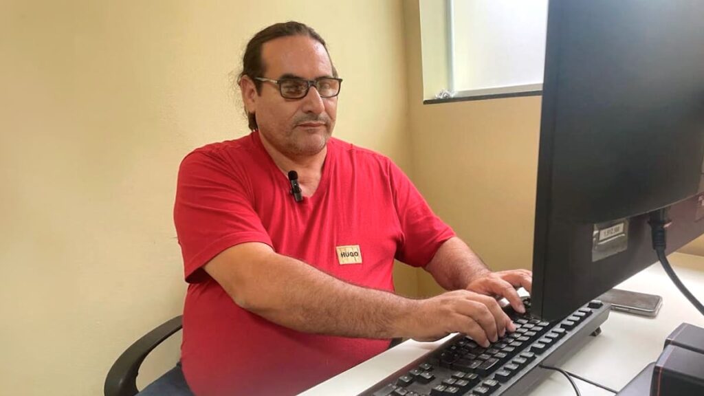 Clodoaldo, um homem usando uma camiseta vermelha, digitando em um computador.