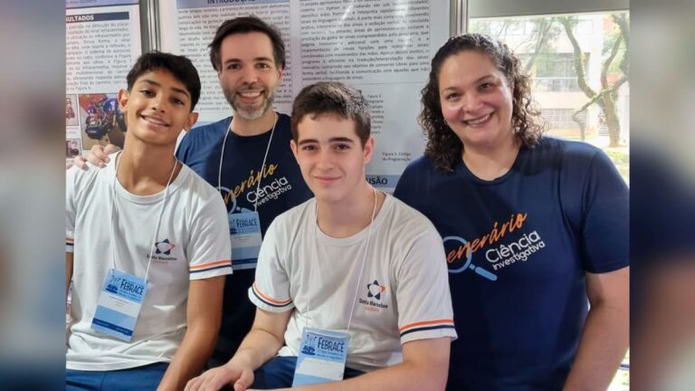 Dois alunos e dois professores posando para uma foto em frente a um pôster do projeto ‘Bastão Tradutor de Libras’, finalista da Febrace 2024.