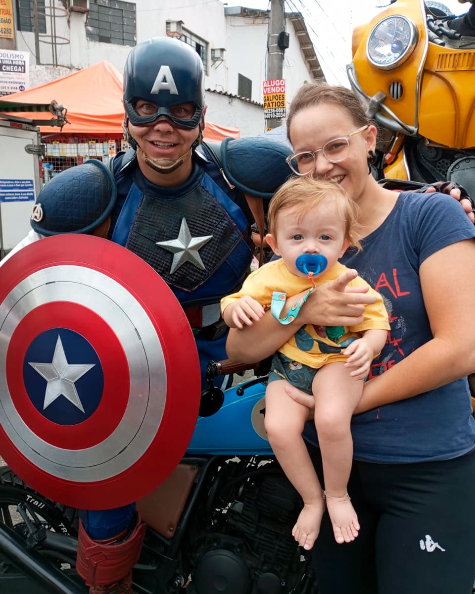 Pessoa fantasiada de Capitão América posando para foto com a mãe Viviane e o Rafinha ao lado de uma motocicleta.