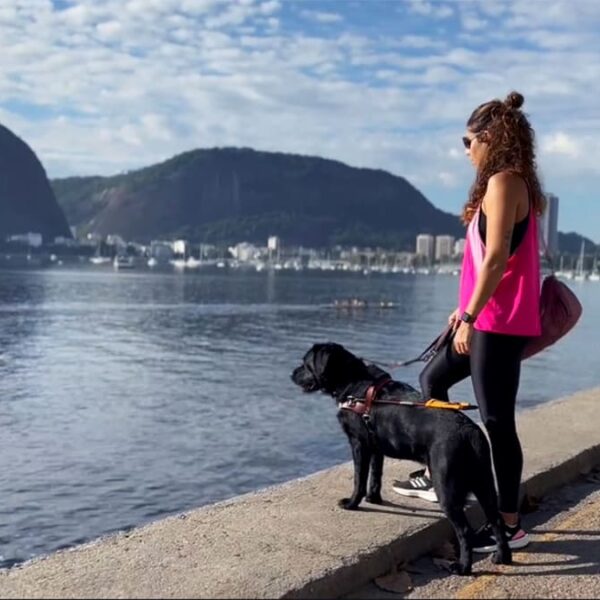 Personagens da segunda temporada série Amor de Bicho, Camila Alves com seu cão-guia Pix, em pé na beira de uma baía, com montanhas e a cidade ao fundo.