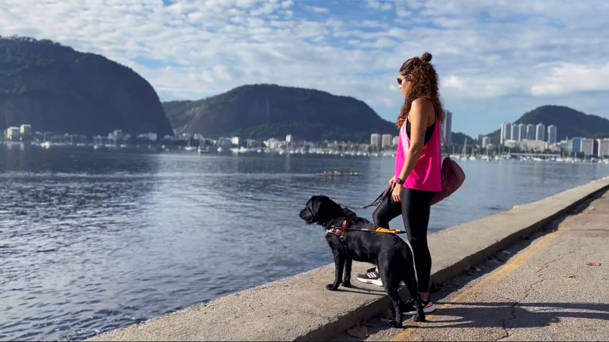 Personagens da segunda temporada série Amor de Bicho, Camila Alves com seu cão-guia Pix, em pé na beira de uma baía, com montanhas e a cidade ao fundo.