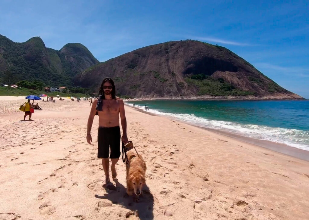 Jonas, um homem cego caminhando com Trevor, seu cão-guia dourado, em uma praia ensolarada com montanhas ao fundo.