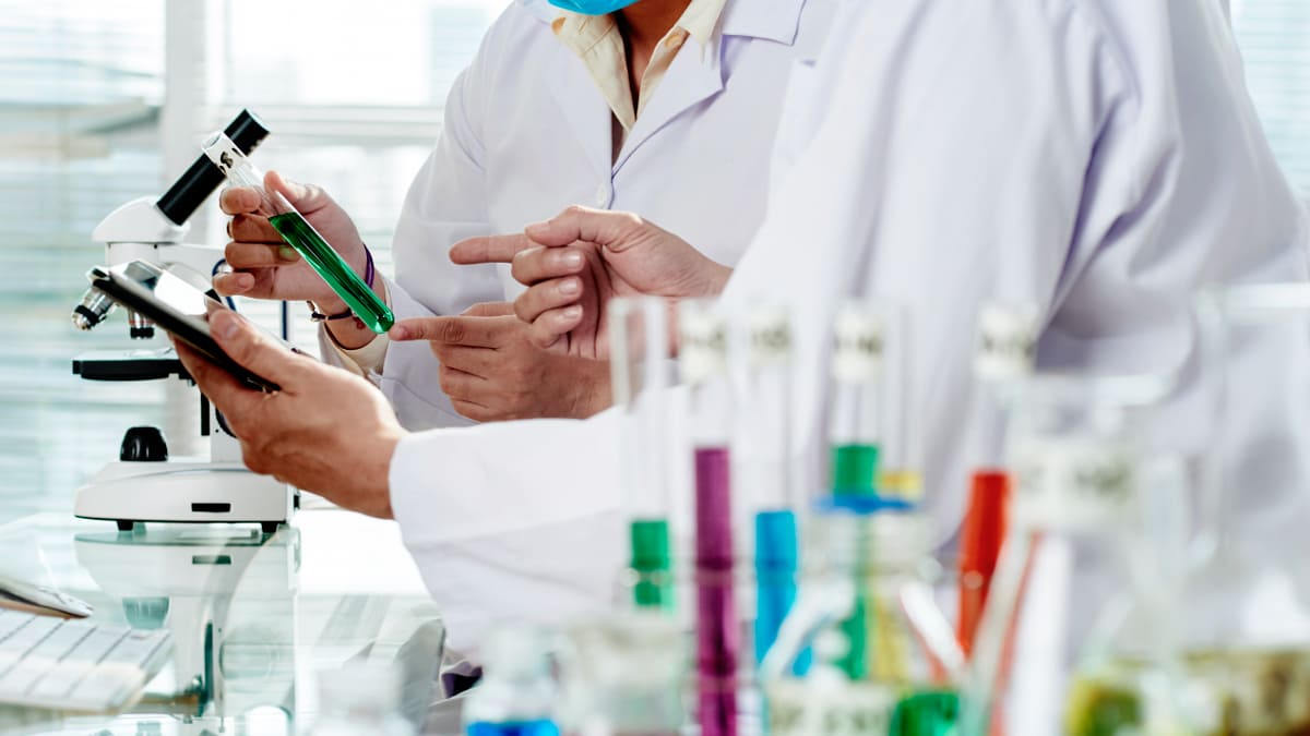 Dois cientistas vestidos com jalecos brancos, conduzindo pesquisa clínica em um laboratório, com um microscópio e tubos de ensaio coloridos no fundo.