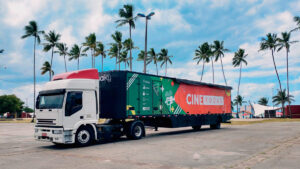 Caminhão branco com carreta colorida do projeto "Cine Rodas" estacionado em um local aberto com palmeiras ao fundo. A lateral exibe imagens e logotipos coloridos, incluindo o título do projeto.