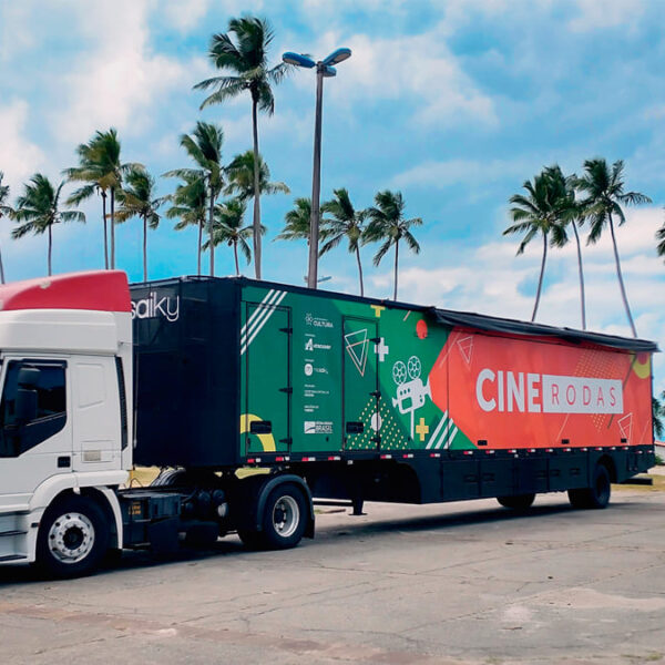 Caminhão branco com carreta colorida do projeto "Cine Rodas" estacionado em um local aberto com palmeiras ao fundo. A lateral exibe imagens e logotipos coloridos, incluindo o título do projeto.