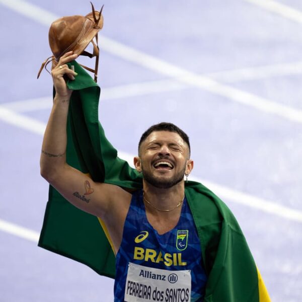 Atleta Petrucio Ferreira celebra o ouro para a chuva de medalhas do Brasil e novo recorde nos 100m do atletismo. O paraibano comemora segurando um chapéu tradicional e envolto na bandeira verde e amarela.