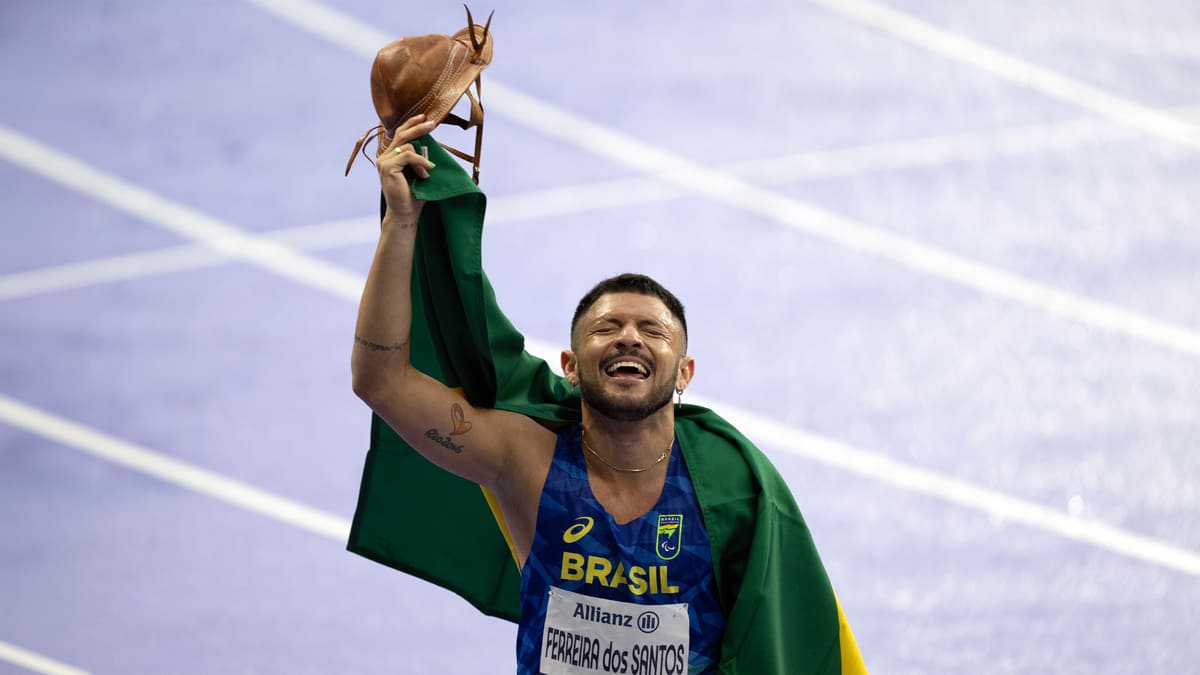 Atleta Petrucio Ferreira celebra o ouro para a chuva de medalhas do Brasil e novo recorde nos 100m do atletismo. O paraibano comemora segurando um chapéu tradicional e envolto na bandeira verde e amarela.