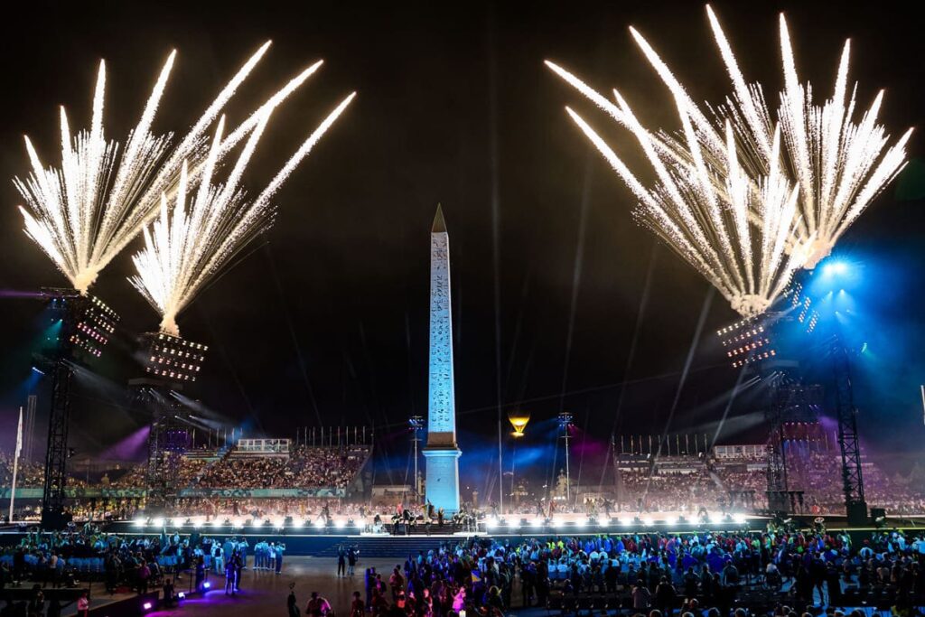 Fogos de artifício explodem no céu noturno ao redor de um grande obelisco iluminado, durante a cerimônia de abertura dos Jogos Paralímpicos de Paris 2024.