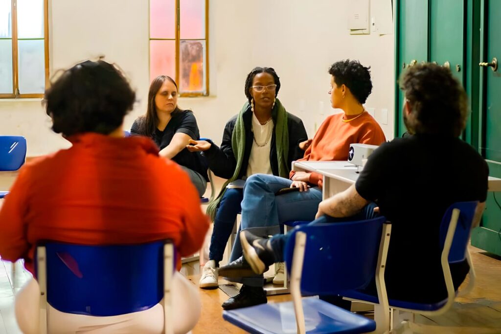 Pessoas participando de uma discussão em grupo durante uma atividade em sala de aula, com foco em uma mulher que fala enquanto os outros escutam atentamente.