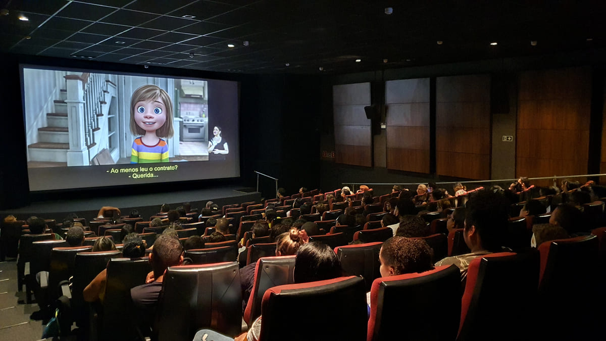 Uma sala de cinema exibe uma cena do filme “Divertida Mente” com a protagonista Riley Andersen, durante o Festival de Cinema Acessível Kids. O público está sentado, assistindo ao filme.