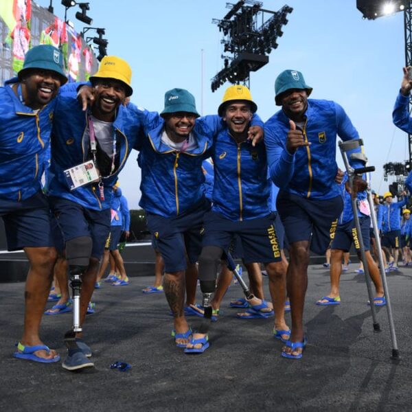 Atletas do Brasil, usando uniformes azuis com "Brasil" escrito, sorriem e em grupo na Abertura da Paralimpíada de Paris 2024. Alguns utilizam próteses ou muletas, todos usando chinelos e chapéus coloridos, comemorando durante o evento esportivo.