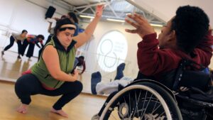 Duas pessoas participam da Oficina DanceAbility, uma delas em uma cadeira de rodas com os braços erguidos e a outra em uma postura de agachamento, ambas em uma coreografia.