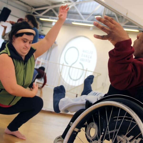 Duas pessoas participam da Oficina DanceAbility, uma delas em uma cadeira de rodas com os braços erguidos e a outra em uma postura de agachamento, ambas em uma coreografia.