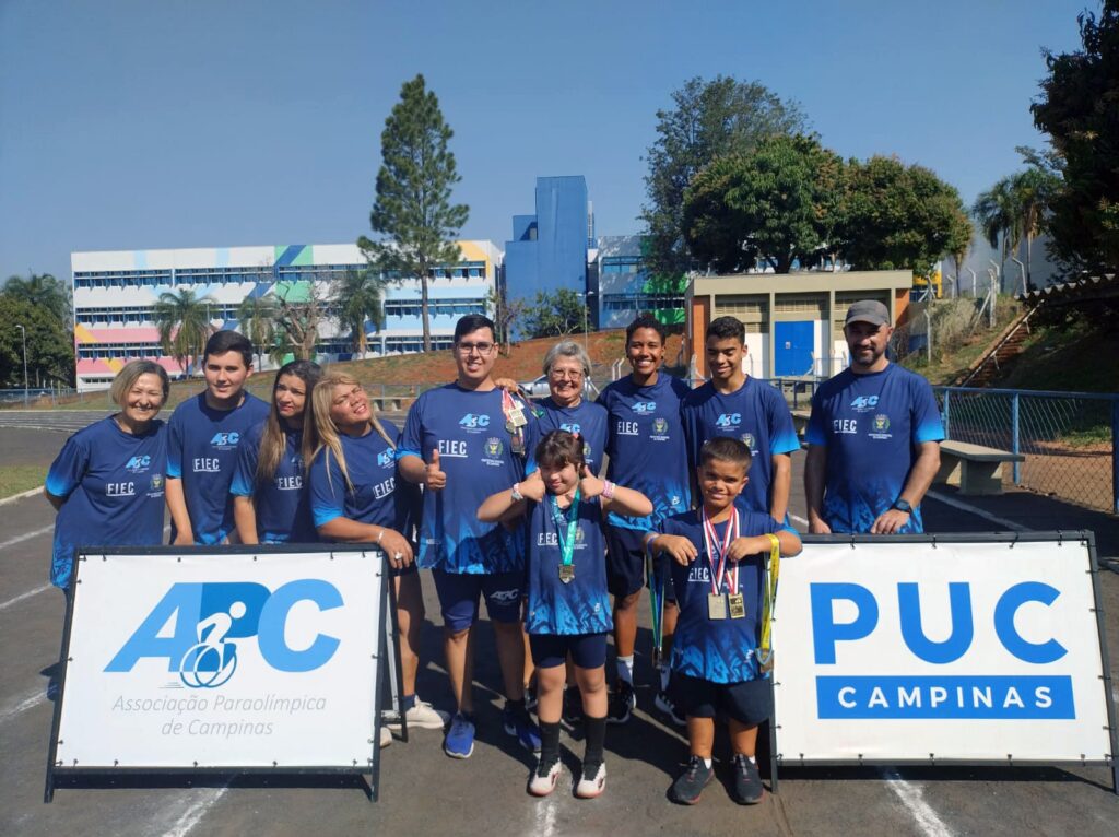 Equipe do projeto "Esportes Sem Barreiras" posando para foto em frente a dois banners com os logotipos da APC e PUC-Campinas.