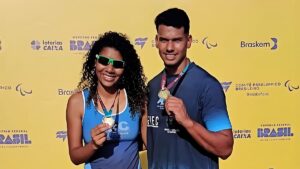 Kétyla e Kesley Teodoro, atletas com deficiência visual, sorrindo e exibindo suas medalhas em frente a um banner amarelo de evento paralímpico.