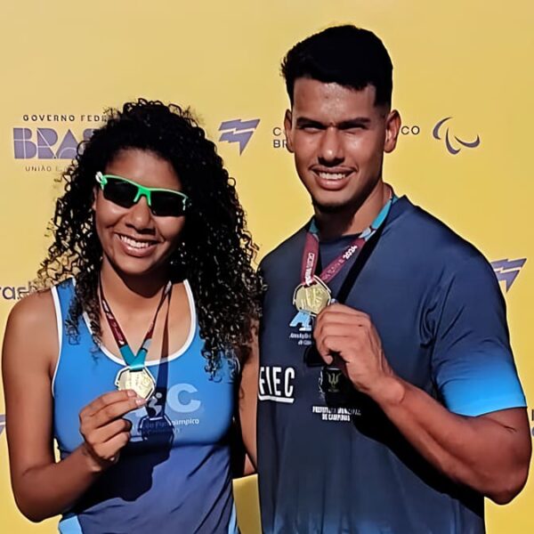 Kétyla e Kesley Teodoro, atletas com deficiência visual, sorrindo e exibindo suas medalhas em frente a um banner amarelo de evento paralímpico.