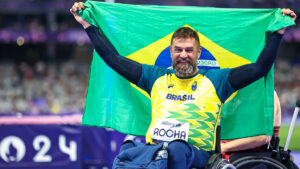 André Rocha, atleta paralímpico em cadeira de rodas, comemora os resultados do Brasil com a bandeira Nacional. Ele veste uniforme nas cores verde e amarelo nos Jogos de Paris 2024.